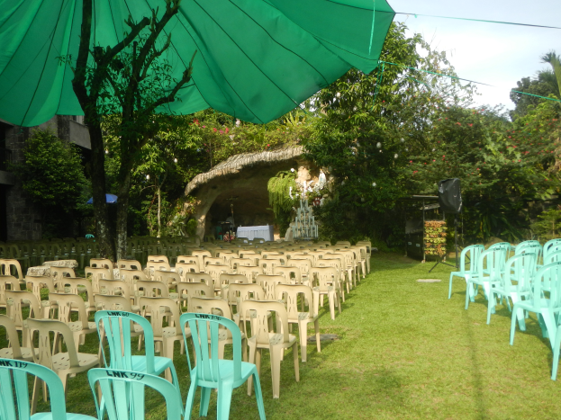 Eine Hochzeitszeremonie in einem Garten mit grünen Stühlen und Schirmen, umgeben von üppiger Vegetation, mit einem Gebäude und einer Hütte im Hintergrund und dem Himmel oben sichtbar.