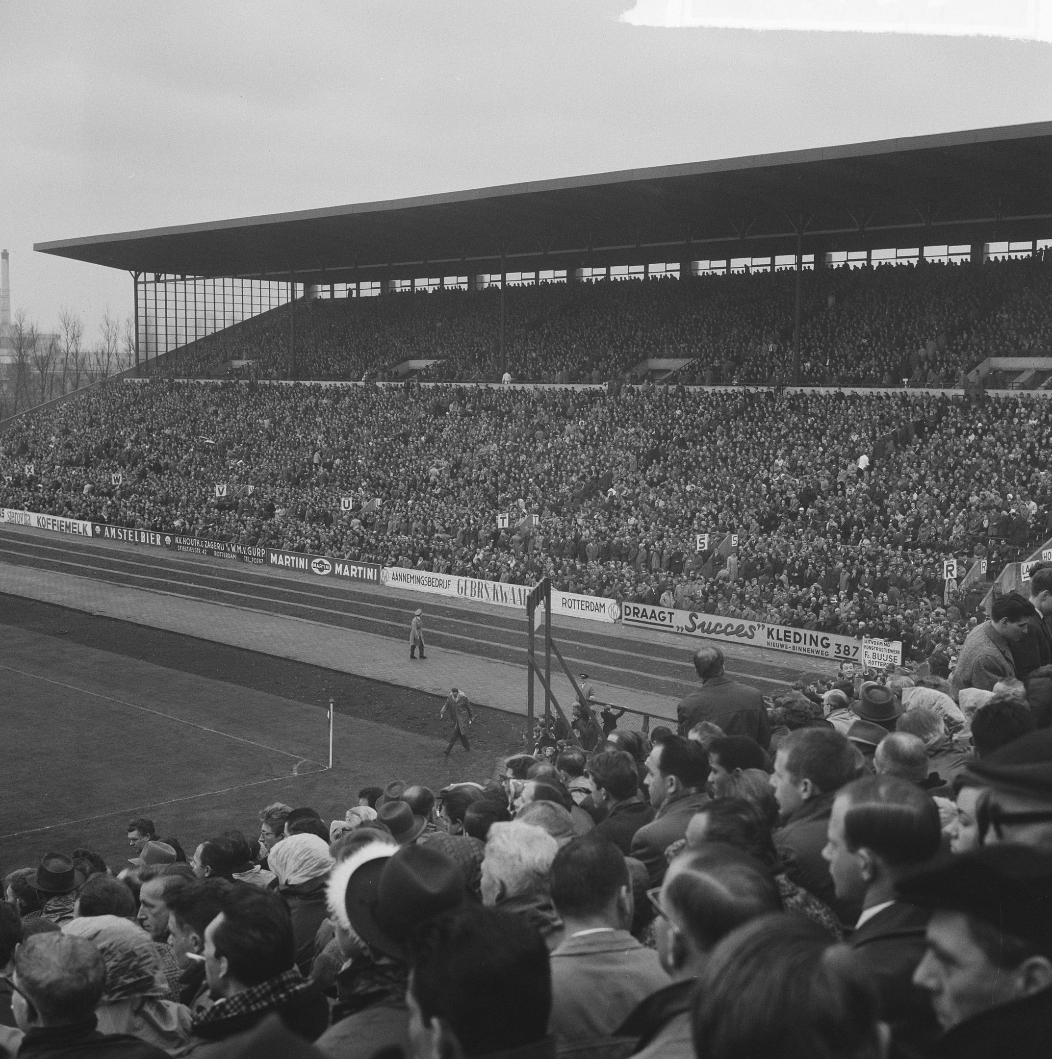 Schwarzes und weißes Foto eines vollen Fußballstadions mit Zuschauern, die stehen und sitzen, Schildern, Pfählen, einem Schuppen, Bäumen, einem Turm und einem bewölkten Himmel.