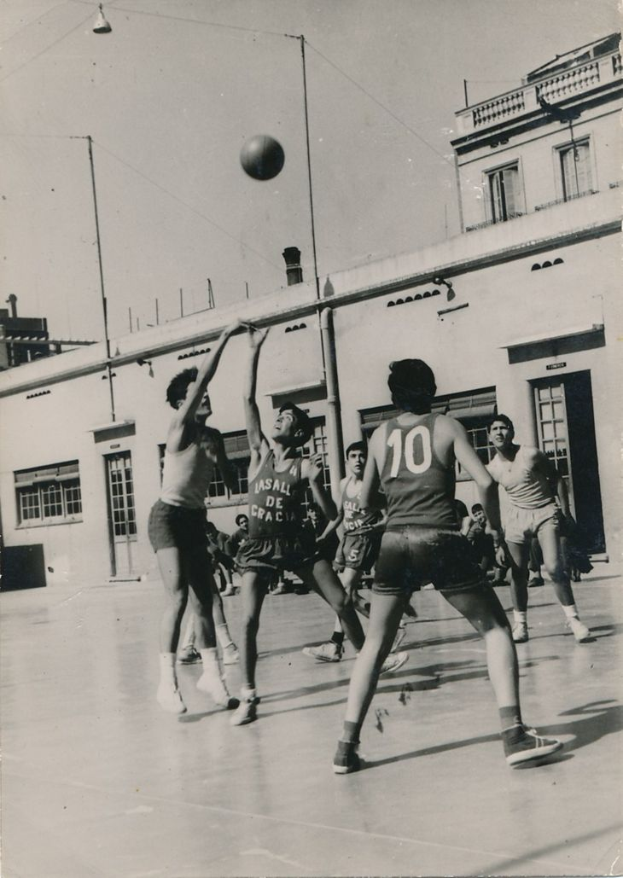 Eine Gruppe von Männern, die Basketball auf einem Platz spielt, mit einem Ball in der Luft und Gebäuden im Hintergrund, dargestellt in Schwarz-Weiß.