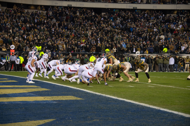 Ein Fußballspiel im Gange mit Spielern auf dem Boden in Sportkleidung und Helmen, von einem Stadion aus mit Zuschauern und Schildern im Hintergrund betrachtet.