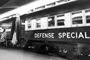 Ein Schwarz-Weiß-Foto eines Zuges mit der Aufschrift "Defense Special", der auf den Schienen steht, mit zwei Fahnen auf der linken Seite und einer Decke darüber.