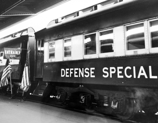 Ein Schwarz-Weiß-Foto eines Zuges mit der Aufschrift "Defense Special", der auf den Schienen steht, mit zwei Fahnen auf der linken Seite und einer Decke darüber.