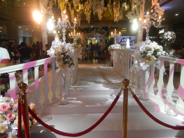 Eine Hochzeitszeremonie in einem Einkaufszentrum mit weißem Teppich, dekorativen Geländern, Blumenarrangements, Kronleuchtern und Deckenbeleuchtung, bei der zahlreiche Gäste anwesend sind, von denen einige Taschen tragen.