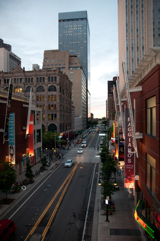 Ein Stadtbild mit einer Straße, flankiert von Gebäuden und Wolkenkratzern, unter einem Himmel.