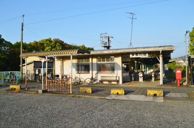 Ein Bahnhof mit vor ihm geparkten Fahrrädern, eine Straße unten, Häuser und Bäume im Hintergrund, Strommasten mit Drähten und einen klaren blauen Himmel.