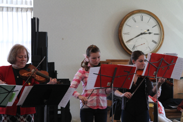 Drei Frauen auf der Bühne mit Geigen stehend und Musikständer vor sich, während eine Gruppe sitzender Frauen im Vordergrund zuhört; eine Uhr hängt an der weißen Wand im Hintergrund, mit einem weißen Vorhang in der linken Ecke.