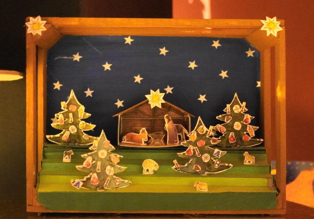 Eine hölzerne Krippe auf einem Tisch, mit einer Lampe links und einer Wand im Hintergrund, dekoriert mit Weihnachtsbäumen, Tieren und einem Haus.