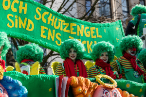 Eine Gruppe von Clowns in bunten Kostümen marschiert durch die Straßen und hält ein Banner hoch, mit Bäumen und einem Gebäude im Hintergrund.