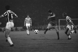 Eine Gruppe von Männern, die Fußball auf einem Feld mit einem Tor im Hintergrund und Zuschauern an der Seitenlinie spielen, dargestellt in Schwarz-Weiß.