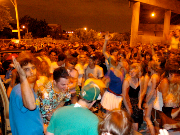 Menschen tanzen im Vordergrund bei einem Konzert mit Lichtern, Pfählen, Bäumen und anderen Objekten im Hintergrund.