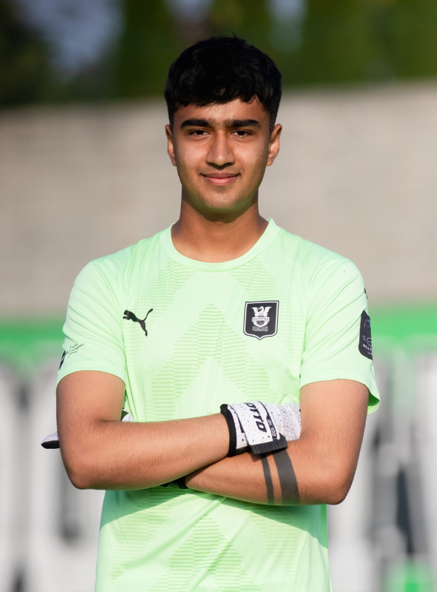 Ein junger Mann in einem grünen Hemd steht auf einem Fußballfeld und hat die Arme verschränkt, er sieht glücklich aus, wahrscheinlich ein Torwart, mit einem unscharfen Hintergrund.
