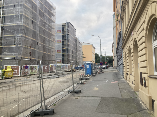 Eine Stra├če mit Geb├Ąuden, Fahrzeugen und einer Baustelle mit Ger├╝st im Vordergrund, unter einem bew├Âlktem Himmel.