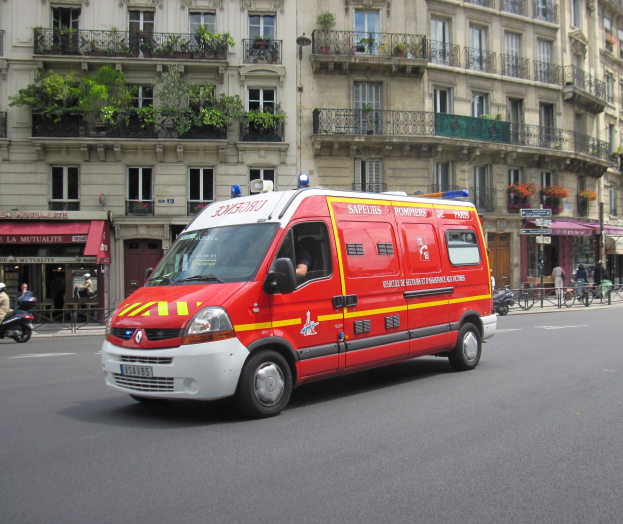 Rettungswagen auf der Straße neben einem Gebäude mit Fenstern, Geländern, Pflanzen und anderen Gegenständen geparkt, mit einigen Menschen und Fahrrädern im Hintergrund.
