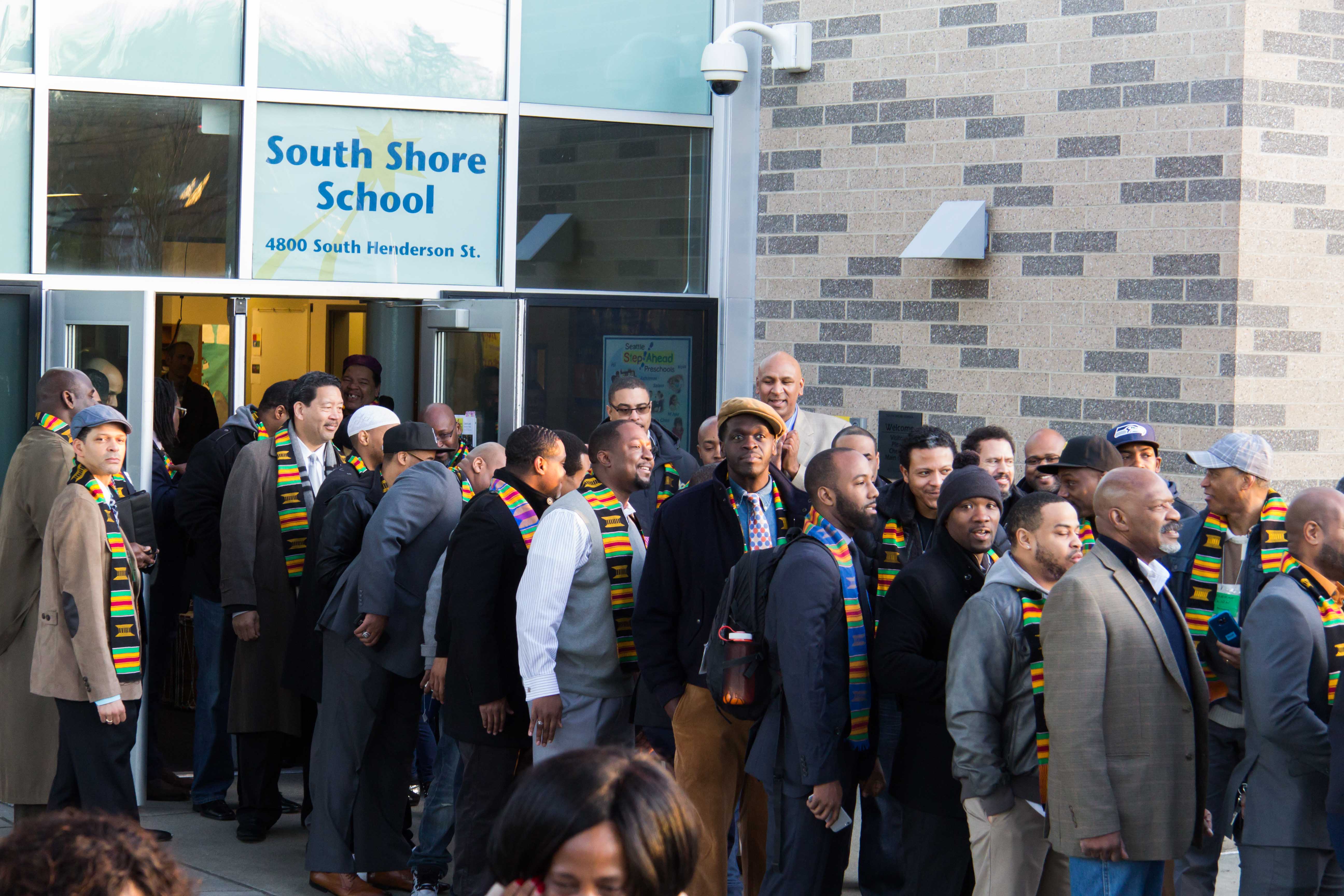 Eine Gruppe von Menschen, einige mit Mützen und Schals, steht vor dem beleuchteten South Shore School-Gebäude mit Glaswänden und -türen und feiert dessen Einweihung, wobei eine Person ein Mobiltelefon hält.