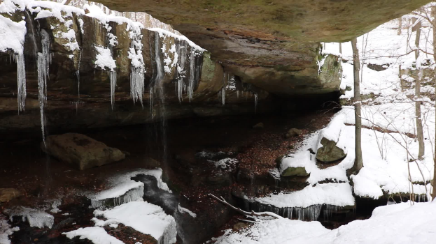 Ein kleiner Wasserfall ergießt sich eine schneebedeckte, felsige Klippe in einem bewaldeten Gebiet, mit Eiszapfen an den Felsen.