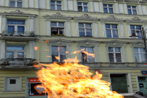Ein großes Feuer brennt vor einem Gebäude mit Fenstern, Türen, einem Lichtmast und einer Tafel mit einer Aufschrift.