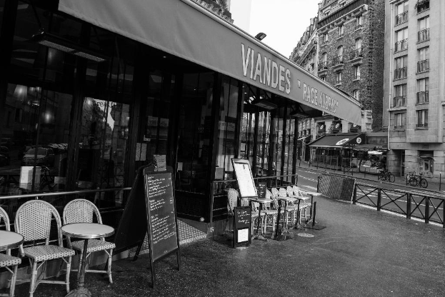 Ein Schwarz-Weiß-Bild einer Straßen Szene mit einem Geschäft und Stühlen in der Mitte, einem Gebäude auf der rechten Seite, einer Straße und Fahrrädern.
