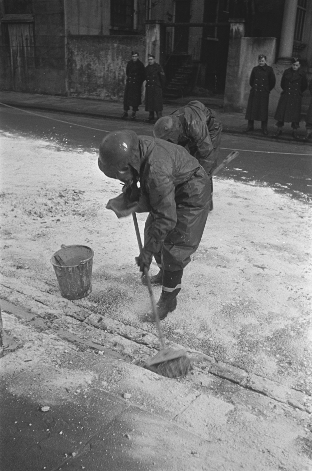 Ein Mann in einem Helm und Handschuhen kehrt Schnee von einem Gehweg mit einer Besen, während eine Gruppe von Menschen auf dem Fußweg in der Nähe steht, mit einem Gebäude mit Fenstern und einer Tür im Hintergrund.