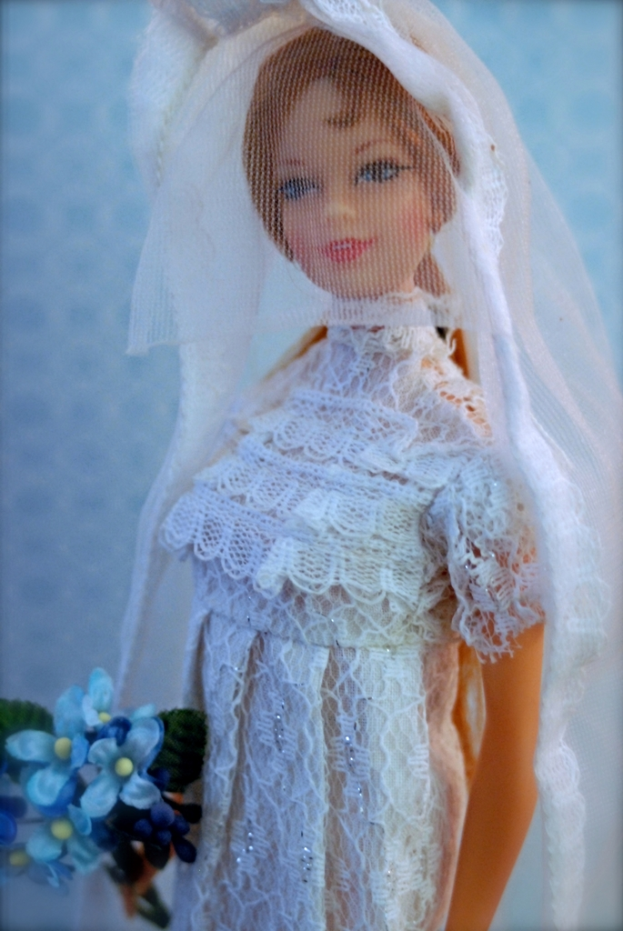 Eine Puppe in einem Kleid, Schleier und mit einem Strauß, mit einem Objekt dahinter.