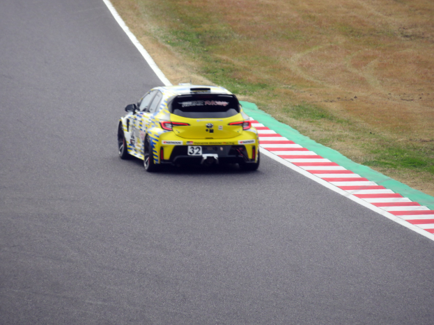 Ein gelber Sportwagen mit Text und Zahlen darauf, der auf einer von Gras gesäumten Rennstrecke fährt.