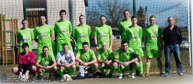 Eine Gruppe von Männern in grünen T-Shirts steht oder hockt auf einem Fußballfeld in der Nähe eines Torpfostens, mit einem Ball davor.