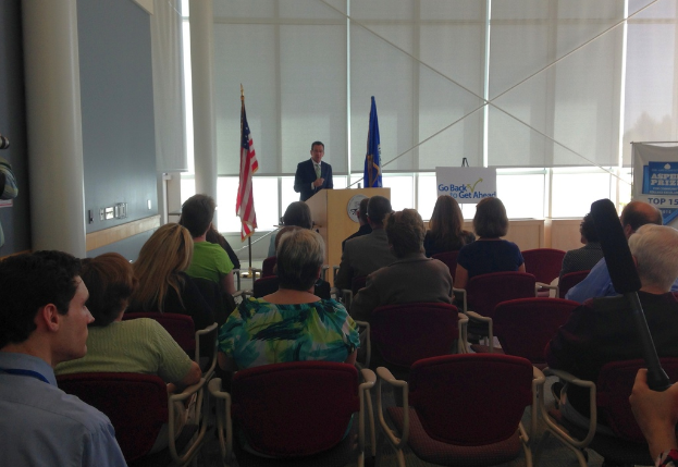 Ein Mann steht an einem Rednerpult und spricht zu einem Publikum, das auf Stühlen sitzt, mit zwei Flaggen daneben und einer Wand, Säulen und Fenstern im Hintergrund.