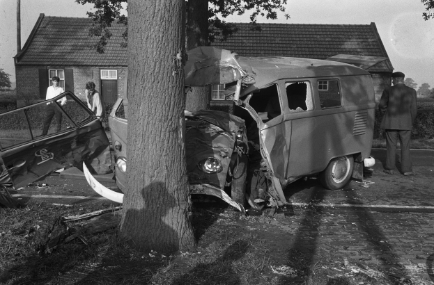 Eine Schwarz-Weiß-Szene eines verunglückten Autos am Straßenrand mit einem Baum im Vordergrund, einigen Menschen in der Nähe und Häusern, Bäumen und einem Mast im Hintergrund.