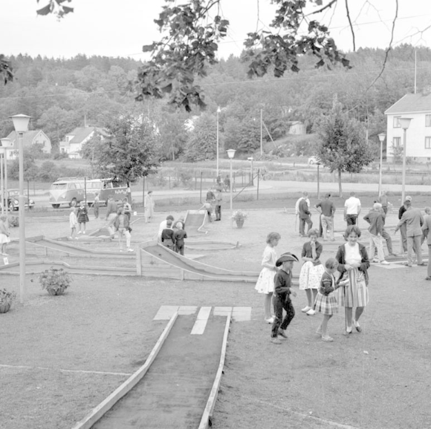 Schwarz-weißes Bild von Menschen, die Minigolf in einem Park mit üppiger Vegetation, Laternenmasten, geparkten Fahrzeugen und Gebäuden im Hintergrund spielen.