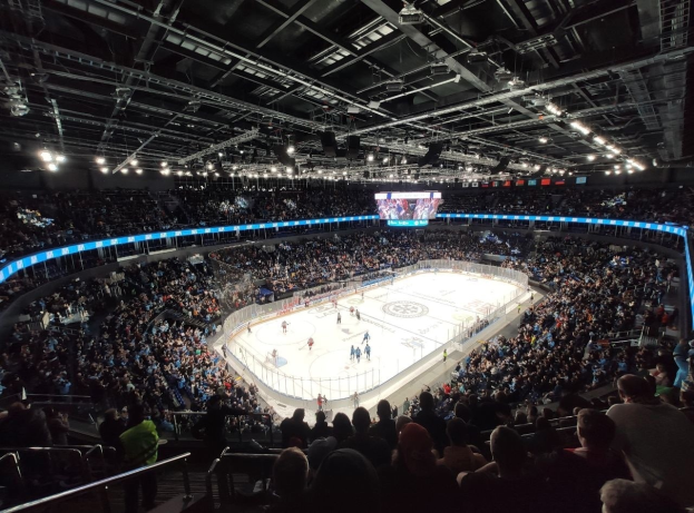 Ein Eishockeyspiel wird im O2 Arena in London gespielt, mit Zuschauern auf den Tribünen und Spielern auf dem Eis, unter hellen Lichtern mit einem Bildschirm an der Spitze des Bildes.