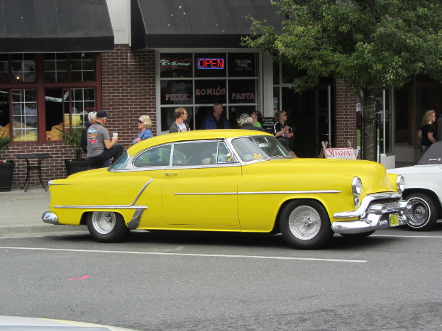 Eine Straßenszene mit mehreren Autos, einigen Fußgängern, Stühlen, einem Baum und einem Gebäude im Hintergrund.