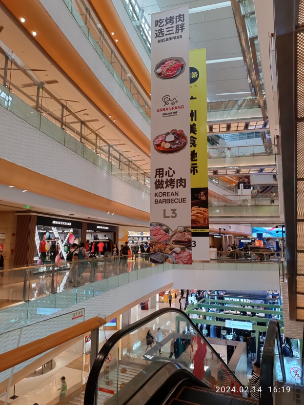 Ein Blick ins Innere eines Einkaufszentrums mit einer Rolltreppe in der Mitte, Menschen, die herumlaufen, einige mit Handtaschen, und Schilder, die von der Decke hängen, mit Geschäften im Hintergrund.