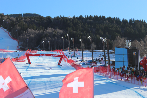 Eine Gruppe von Menschen steht auf einer schneebedeckten Anhöhe mit roten Fahnen im Vordergrund, Laternen, Bäumen und einem klaren blauen Himmel im Hintergrund und einem großen Bildschirm auf der rechten Seite.