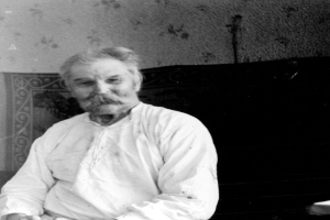 Ein alter Mann mit Schnurrbart sitzt in einem Stuhl vor einer Wand, trägt ein weißes Hemd und hat einen ernsten Gesichtsausdruck, dargestellt auf einem Schwarz-Weiß-Foto.