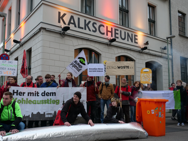 Menschen bei einer Demonstration vor einem Gebäude mit Schildern und Plakaten, zwei Personen sitzen im Vordergrund und ein Müllcontainer ist auf der rechten Seite zu sehen.