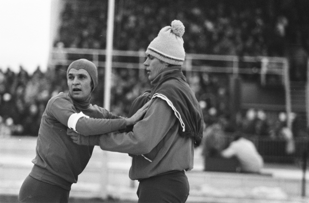 Zwei Männer in Jacken und Mützen auf einem Feld kämpfend mit unscharfen Zuschauern, Pfählen und einem Zaun im Hintergrund, in Schwarz-Weiß.