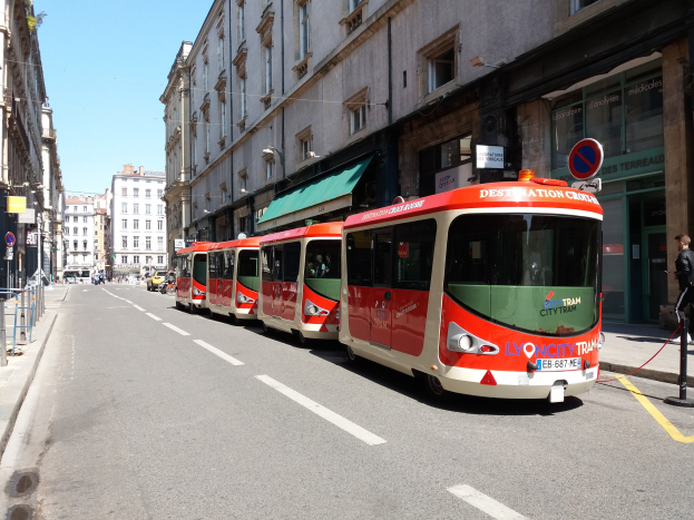 Eine Reihe roter und weißer Busse, die entlang einer Straße in Lyon, Frankreich, geparkt sind, mit Menschen, die auf dem benachbarten Gehweg gehen und Gebäuden, die die Straße säumen.