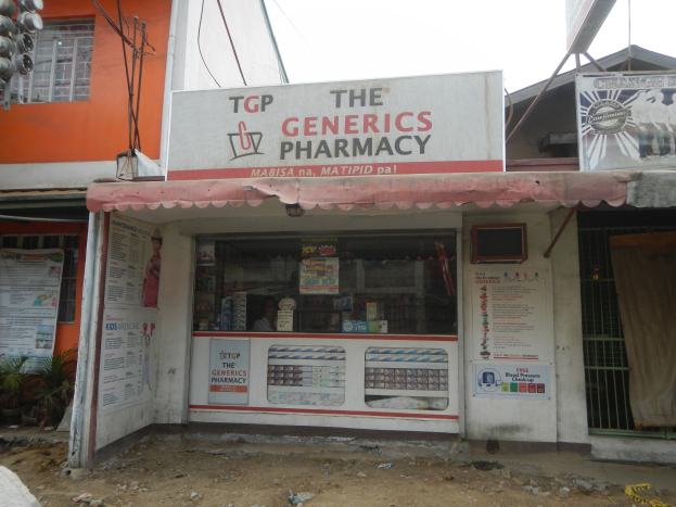 Apothekenfront mit einem "The Generics Pharmacy"-Schild, umgeben von Gebäuden, Strommasten und Fahrzeugen unter einem klaren blauen Himmel.