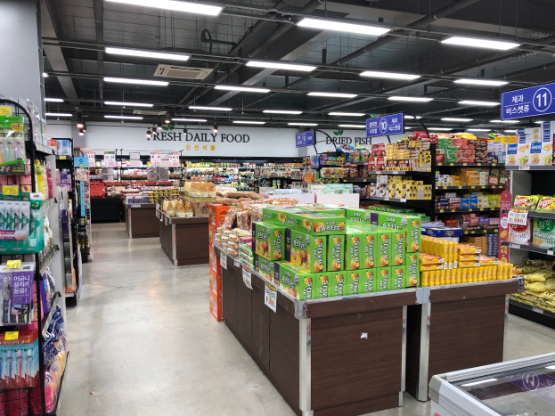 Ein Supermarktgang in Seoul, Südkorea, der verschiedene Obst- und Gemüsesorten sowie andere Lebensmittel auf Regalen zeigt, mit Deckenbeleuchtung und Informationsschildern.