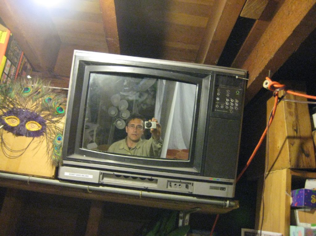 Ein Fernsehbildschirm mit einer Person, umgeben von Pfauenfedern, Büchern und Kisten.