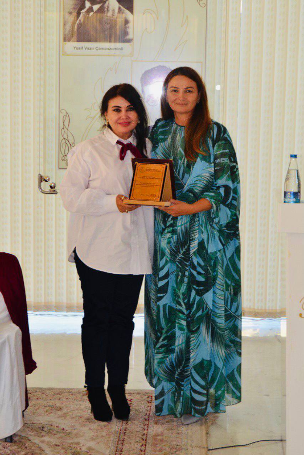 Zwei Frauen stehen nebeneinander und lächeln auf einer Preisverleihung; die Frau links hält einen Preis, während die Frau rechts neben einem Rednerpult mit einer Wasserflasche darauf steht, mit Vorhängen und einem Foto im Hintergrund.