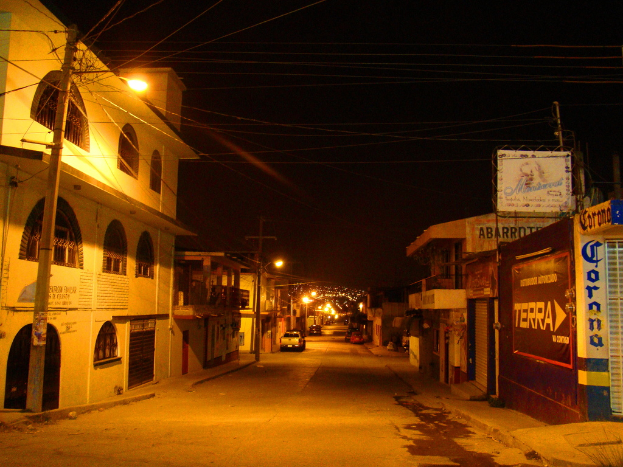 Eine Straßenszene mit Gebäuden und Strommasten auf beiden Seiten, einigen parkenden Fahrzeugen und einer Straße in der Mitte.