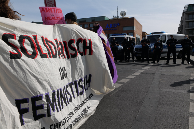 Eine Gruppe von Menschen marschiert auf einer Straße, die ein Banner mit der Aufschrift 'Solidarität und Feminismus' trägt, mit parkenden Fahrzeugen auf der Seite und Gebäuden im Hintergrund unter einem klaren blauen Himmel.