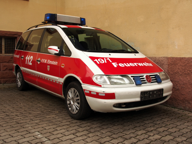 Ein roter und weißer Minivan mit Notarztkennzeichen vor einem Fensterfront-Gebäude geparkt.