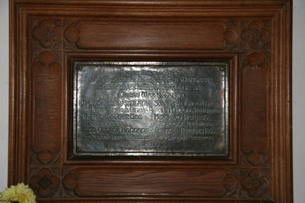 Eine Gedenktafel mit Text ist an einer braunen Wand oder einer hölzernen Tür angebracht, mit einem gelben Objekt, das einer Blumenvase ähnelt, in der linken unteren Ecke.