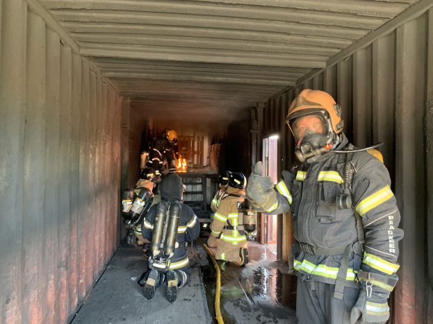 Feuerwehrleute in Schutzausrüstung, darunter Helme und Sauerstoffflaschen, arbeiten innerhalb eines Seecontainers mit einem Feuerlöscher im Hintergrund.
