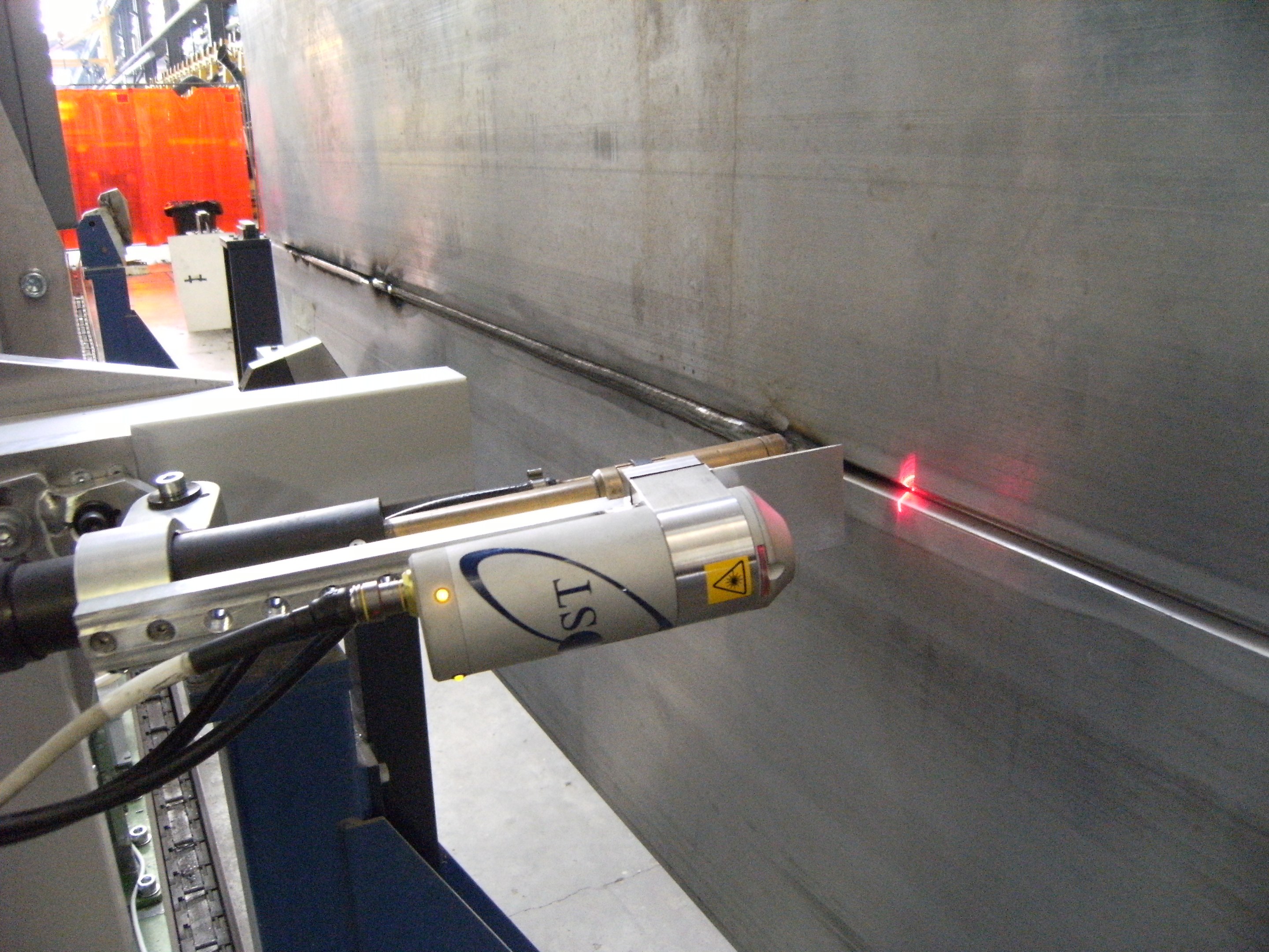 Heller Laserstrahl aus einer Schneidemaschine, der ein Fabrikinnere mit einer Wand auf der rechten Seite und Hintergrundobjekten beleuchtet.