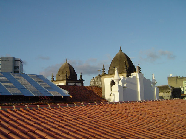 Eine Stadtansicht mit mehreren Gebäuden im Vordergrund unter einem klaren blauen Himmel, die Solarpanels auf dem Dach eines Gebäudes zeigen.
