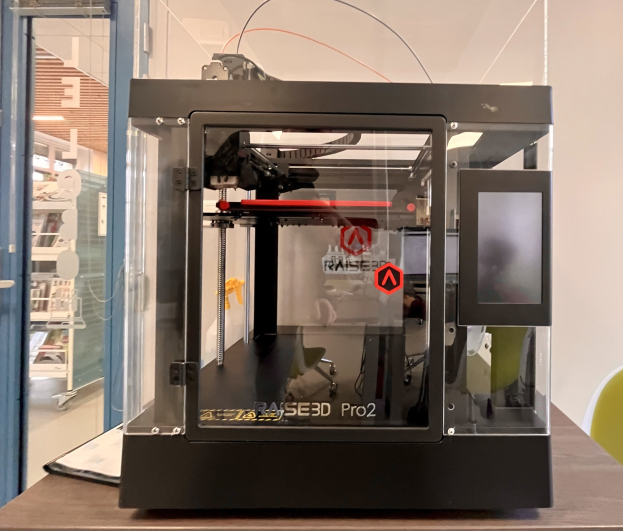 3D-Drucker auf einem Holztisch mit einer Glastür links und einem Stuhl rechts, Regalen mit Gegenständen im Hintergrund.