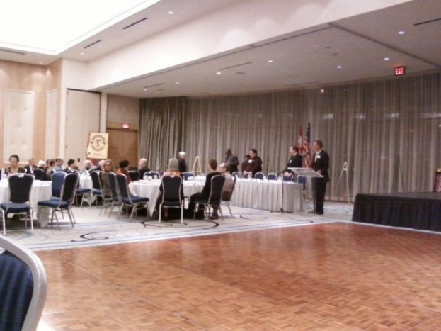 Eine Gruppe von Menschen sitzt und steht in einem Raum mit Flaggen, Lichtern, Schildern und Vorhängen im Hintergrund.