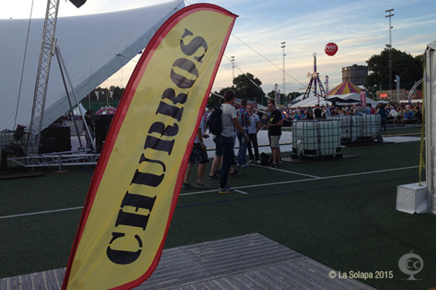 Eine lebhafte Karnevalsszene mit Zelten, einem zentralen Mast, Menschen, Bäumen, Strommasten, einem Himmel, Straßenbahnen auf der Straße und einem Eisenständer auf der linken Seite.